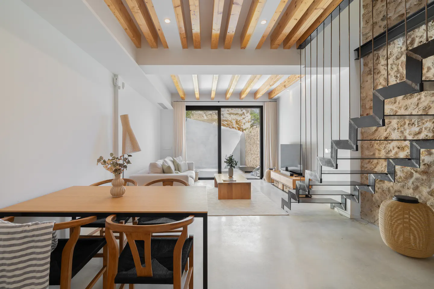 Bright, modern living room with wood-beamed ceiling, stone wall, and metal staircase. Table and chairs in foreground, sofa and TV in background.