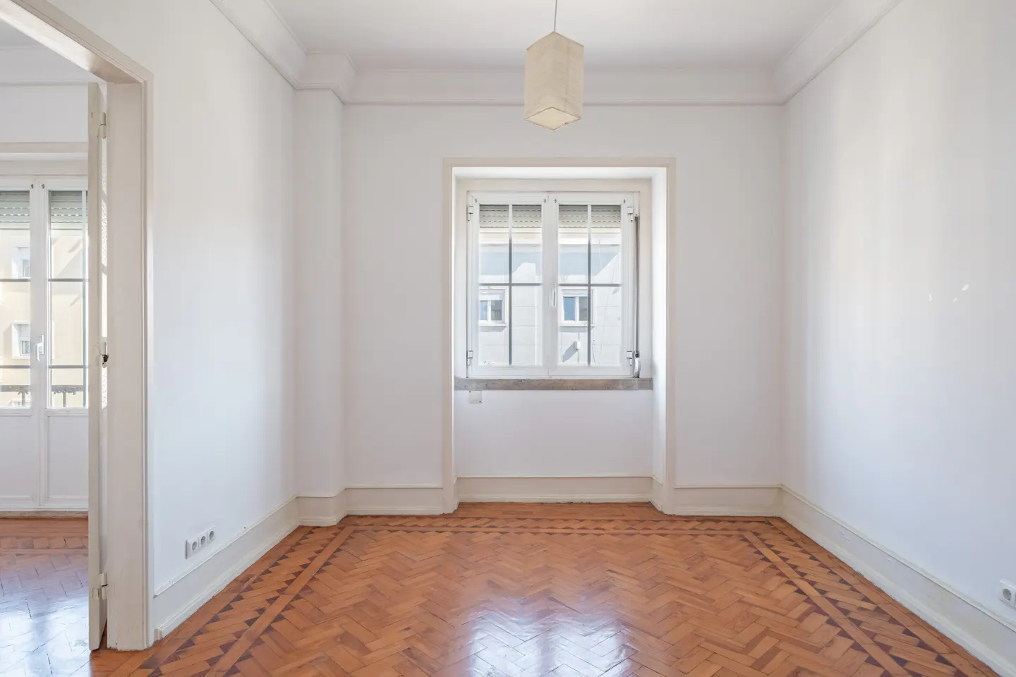 Empty room with white walls, a central window, and polished herringbone wood flooring.
