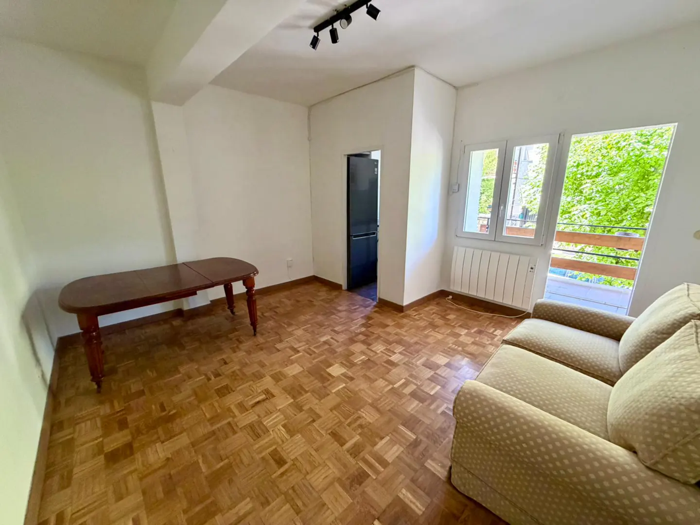 Bright living room with parquet floors, white walls, a brown table, and a beige sofa near a window with a green view.