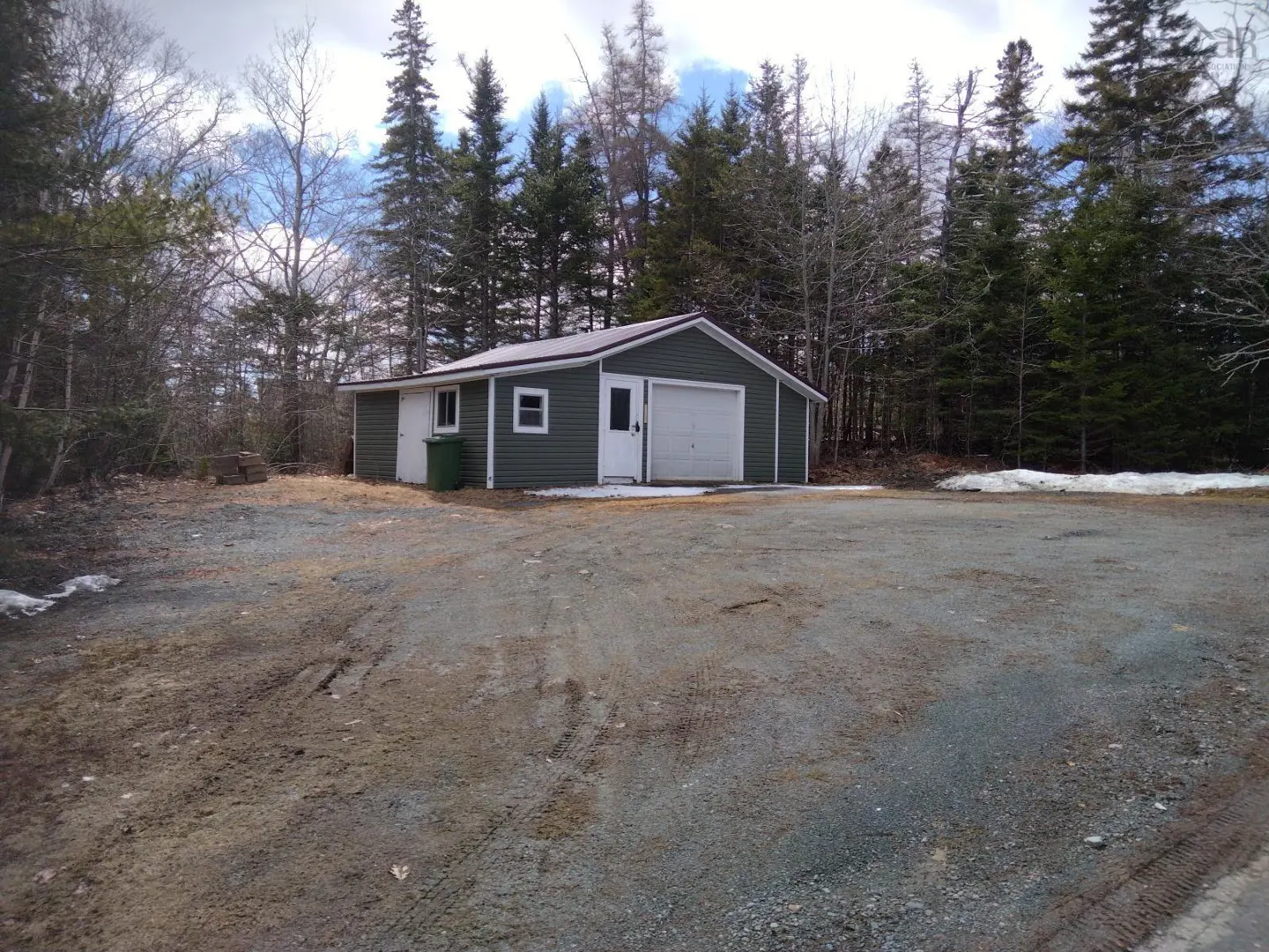 A one-story, green garage with a white door and a gravel driveway. Trees are in the background.