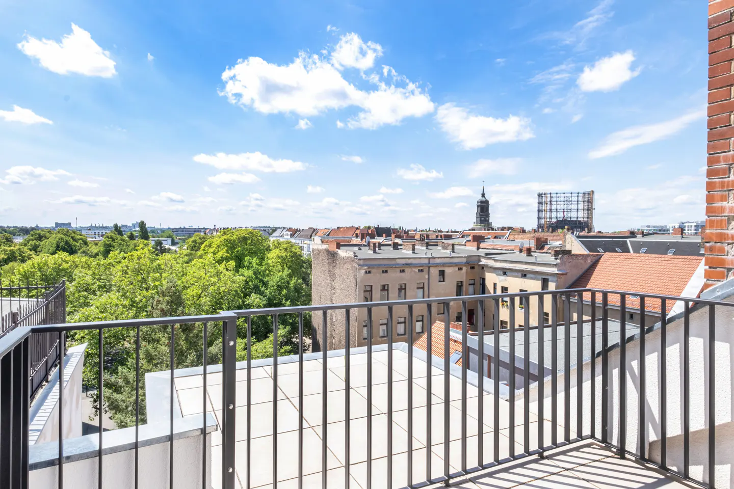 View from a balcony with black railings overlooking a European city with red rooftops, green trees, and a blue sky with white clouds.