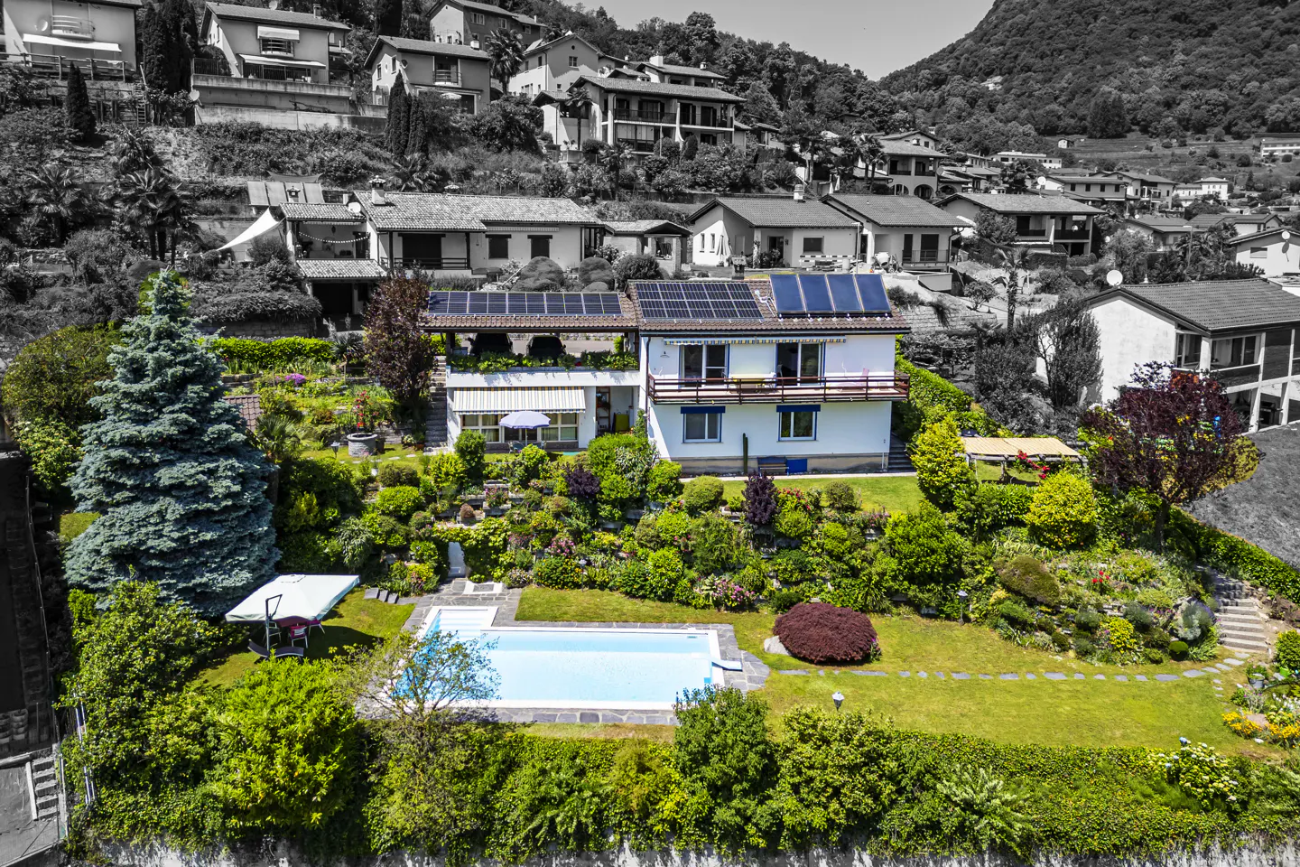 Aerial view of a white two-story house with solar panels, a swimming pool, and lush gardens on a hillside.
