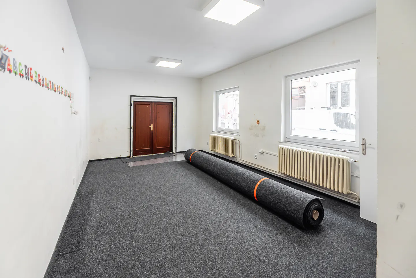 Empty room with gray carpet, white walls, and two windows. A roll of carpet lies on the floor. Brown double doors are in the background.