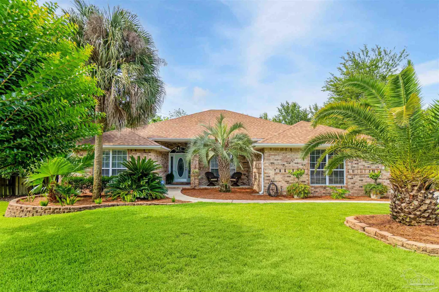 Single-story brick house with a tan roof, lush green lawn, palm trees, and a front porch with two chairs.