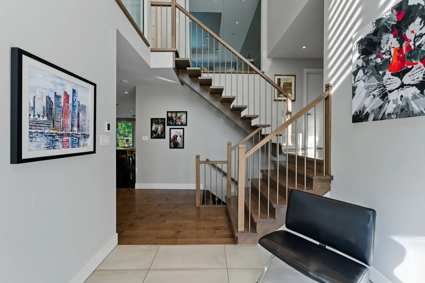 A modern home foyer with a wooden staircase, artwork, and a black bench. Sunlight streams through the window.
