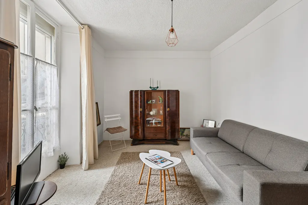 Bright living room with white walls, a gray sofa, and a brown wooden cabinet. A white table sits on a beige rug.