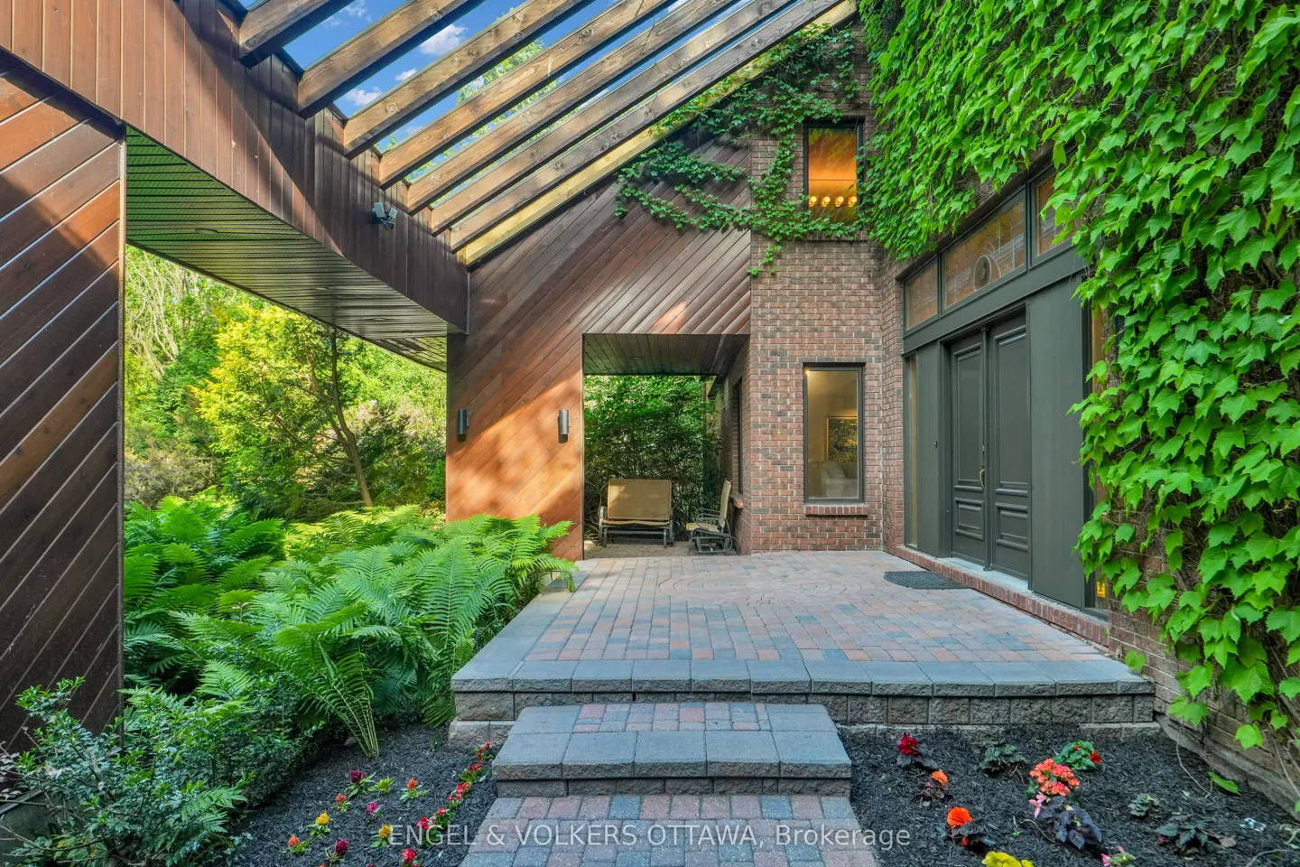 A brick home entrance with a wood-slat pergola, lush green ivy, a stone patio, and a garden with ferns and flowers.