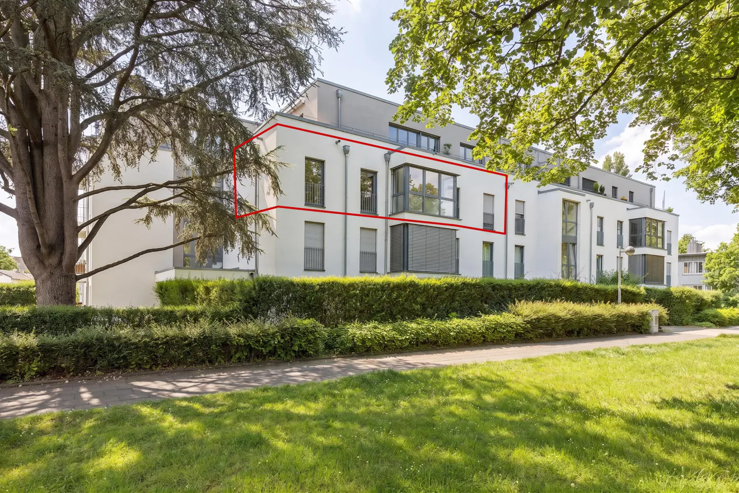Modern white apartment building with a red outline highlighting a specific unit, surrounded by trees and a green lawn.