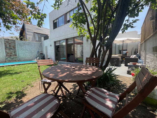 Outdoor patio with a round wooden table and chairs on a green lawn, a pool, and a white two-story house in the background.