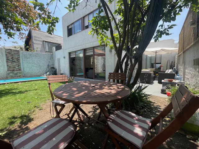 Outdoor patio with a round wooden table and chairs on a green lawn, a pool, and a white two-story house in the background.