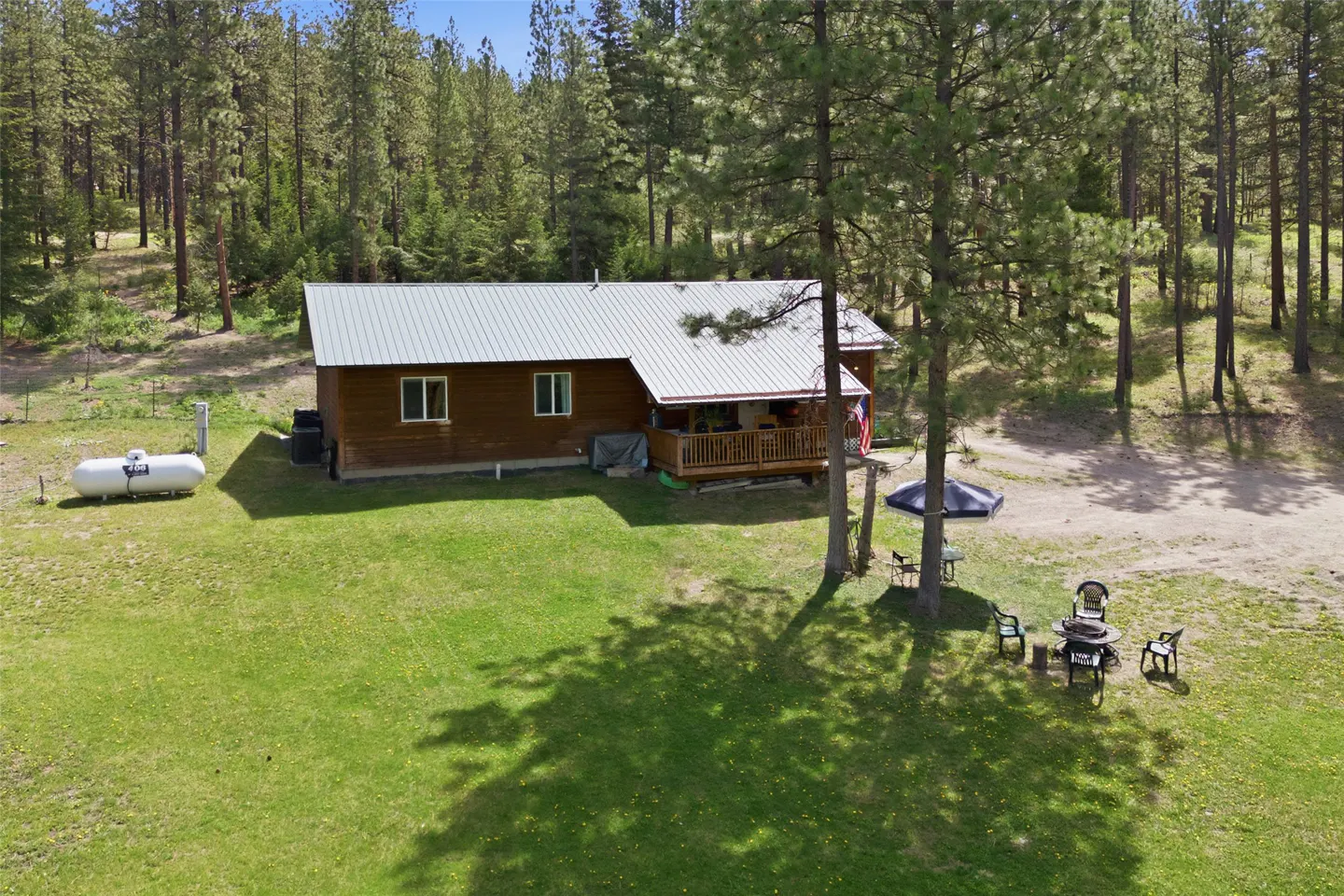 Exterior view of a brown cabin with a metal roof, surrounded by green grass and tall trees. A propane tank sits near the house, and a patio set is visible in the yard.
