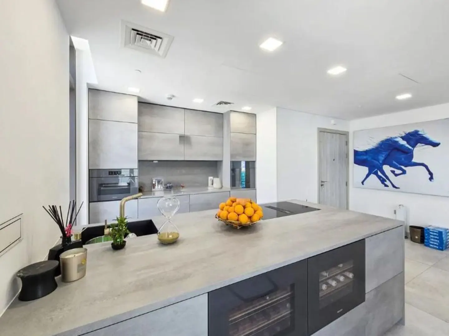 Modern kitchen with gray cabinets, a large island with a bowl of oranges, and a blue horse painting on the wall.