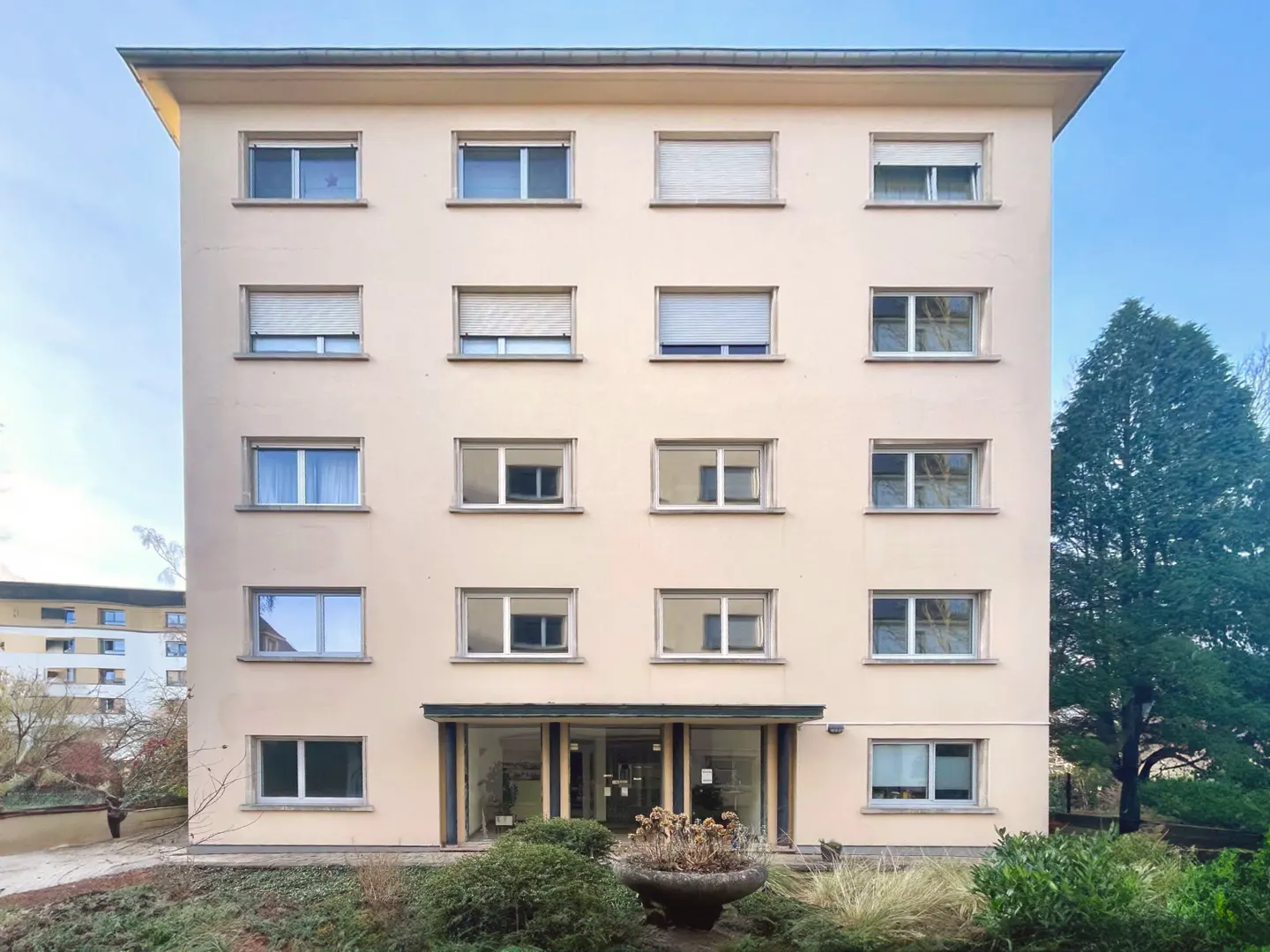 Four-story apartment building with a flat roof, beige facade, and symmetrical window arrangement. Greenery surrounds the entrance.