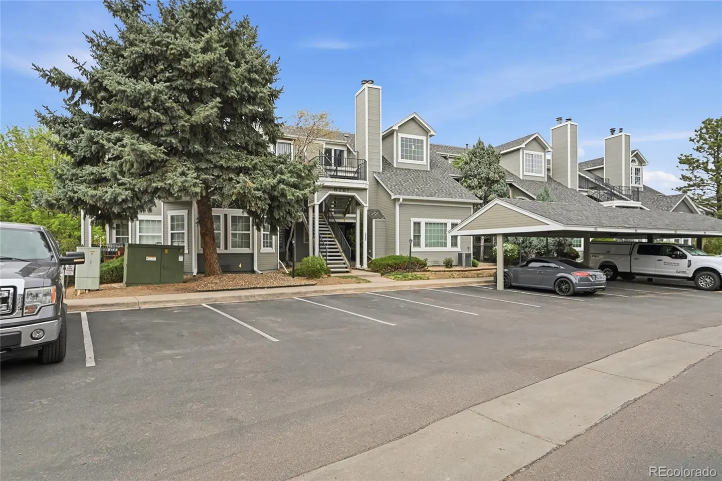 Exterior view of a gray apartment building with a parking lot and cars under a carport.