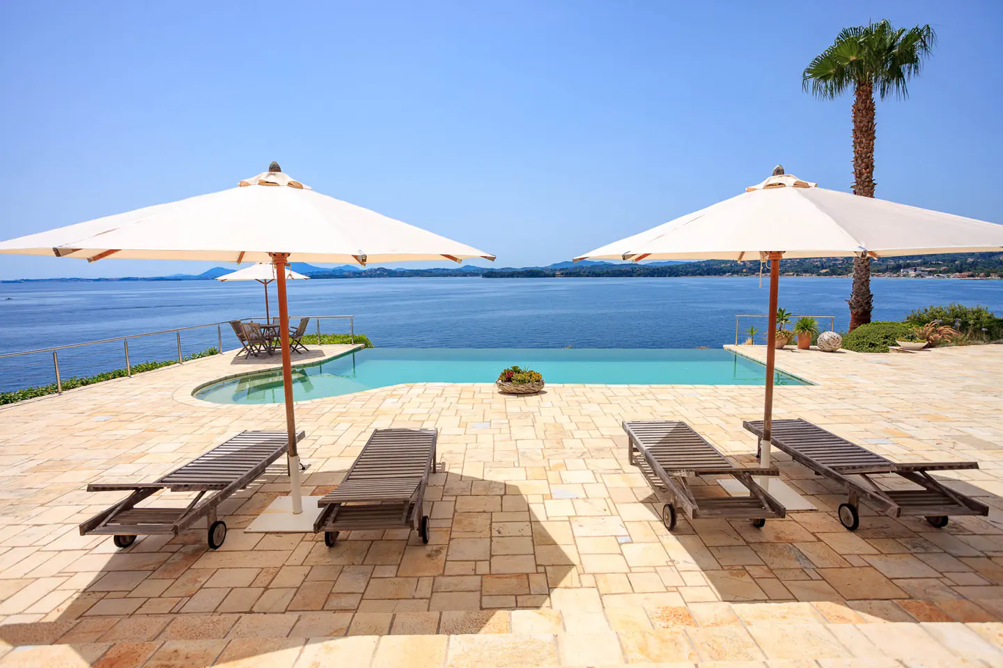 Luxury villa patio with lounge chairs, umbrellas, and infinity pool overlooking the ocean on a sunny day.