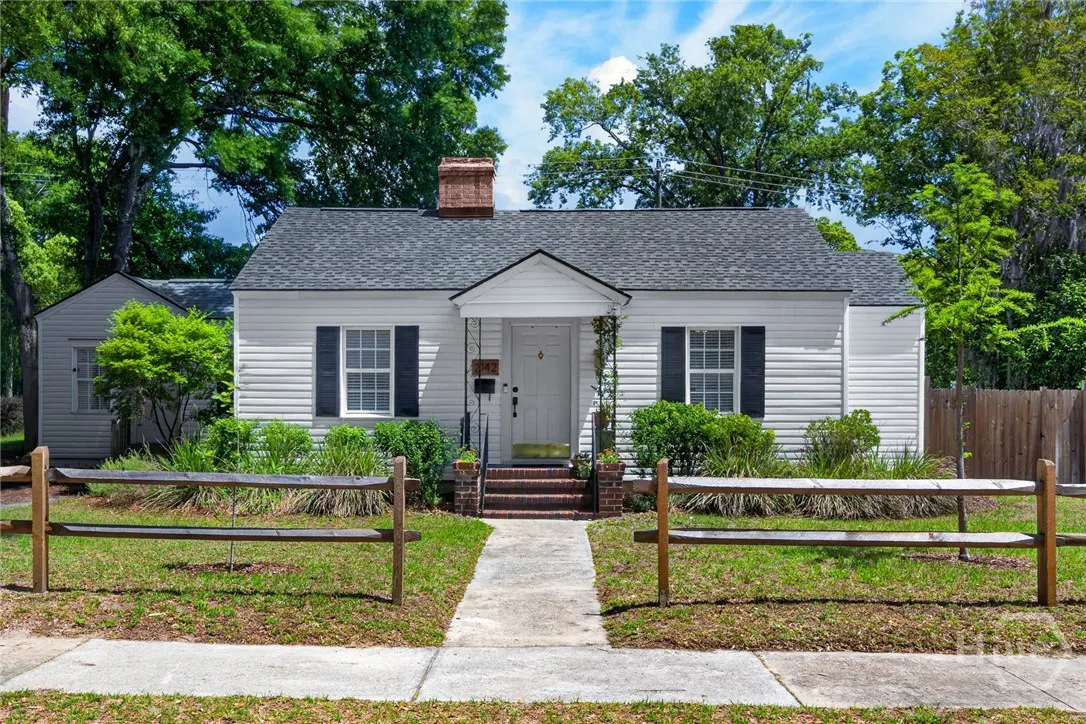 A white, one-story house with black shutters and a gray roof, surrounded by green trees and a wooden fence.