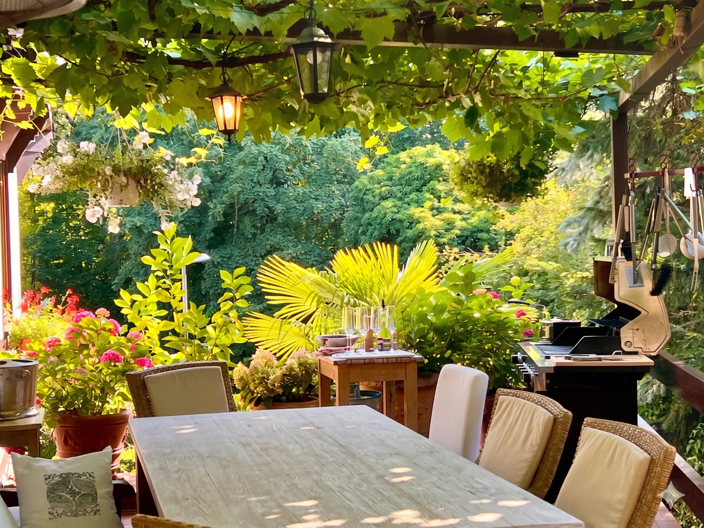 Outdoor dining area with a wooden table, wicker chairs, and lush greenery. A grill and hanging lights add to the ambiance.