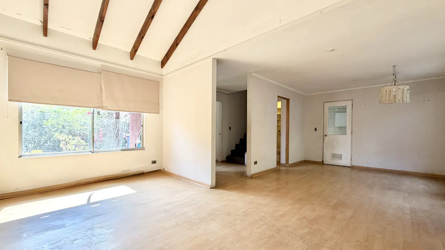 Empty living room with light wood floors, white walls, and a window with beige blinds. Brown wooden beams accent the ceiling.