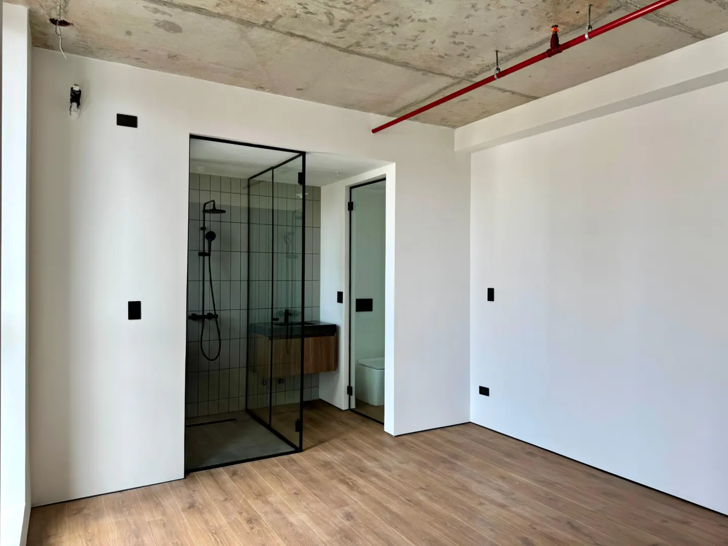 A modern bathroom with a glass shower, wood floors, and white walls. A red pipe runs across the concrete ceiling.