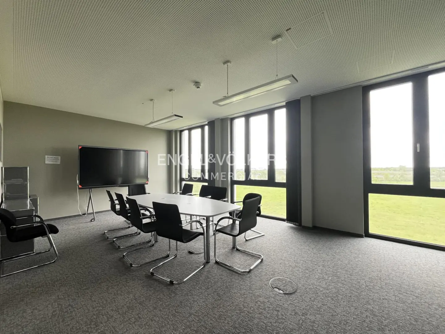A bright conference room with a white table, black chairs, a large screen, and big windows.