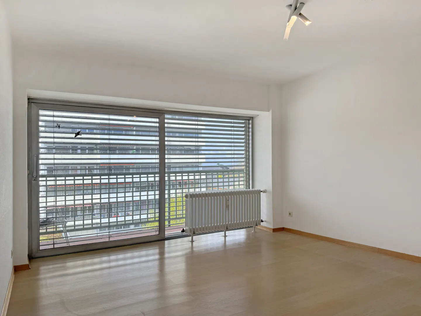 Bright, empty room with wood floors, white walls, and a large window with blinds. A radiator sits below the window. A modern light fixture is on the ceiling.