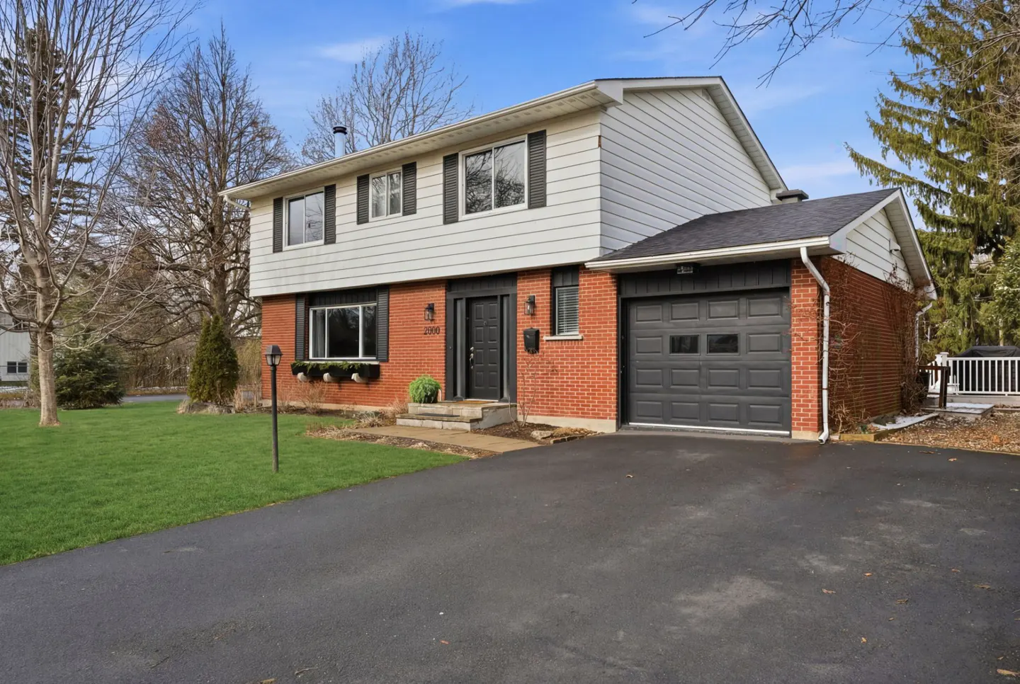 Two-story house with red brick and white siding, black shutters, and a black garage door. A green lawn and asphalt driveway are in front.