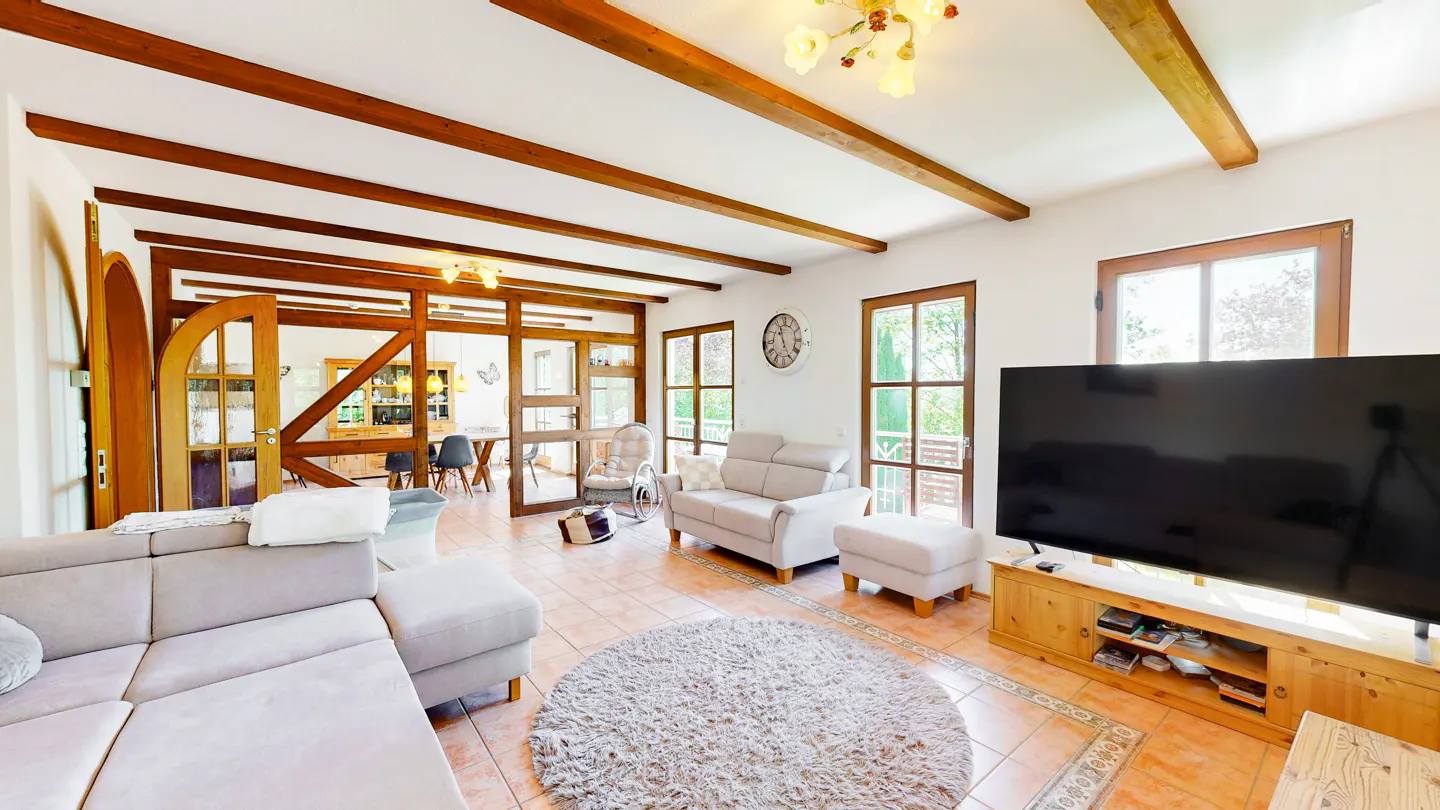 Bright living room with exposed wood beams, tile floor, and gray sofas. A large TV sits on a wooden console.