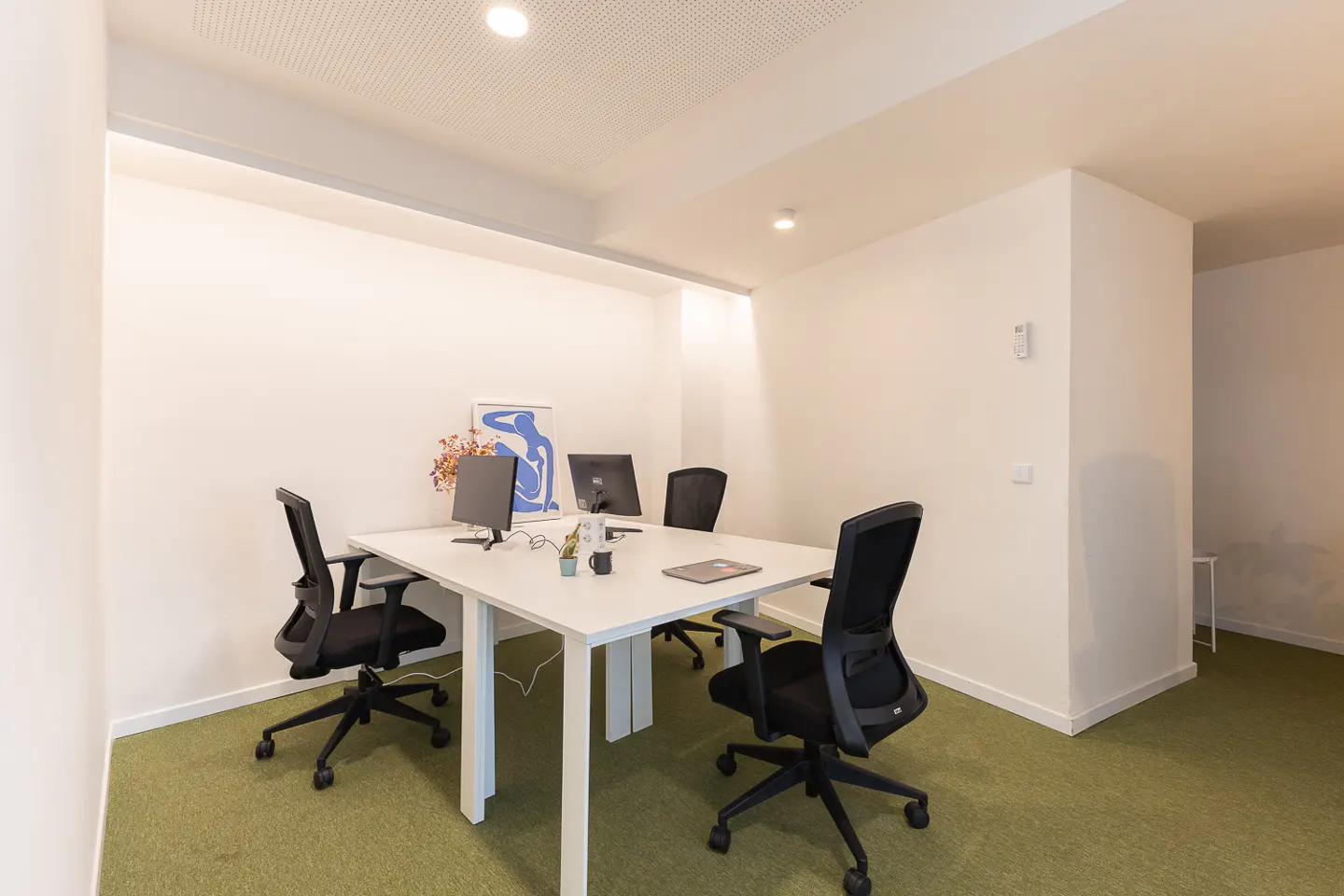Modern office space with a white desk, two computer monitors, and black ergonomic chairs on a green carpeted floor.