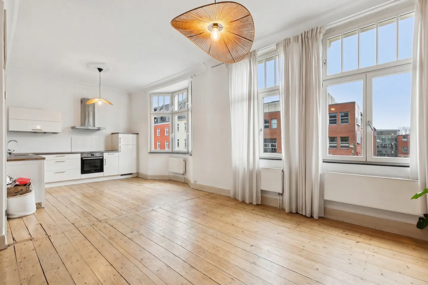 Bright, open apartment with wood floors, white walls, and large windows with white curtains. A modern kitchen is visible in the background.