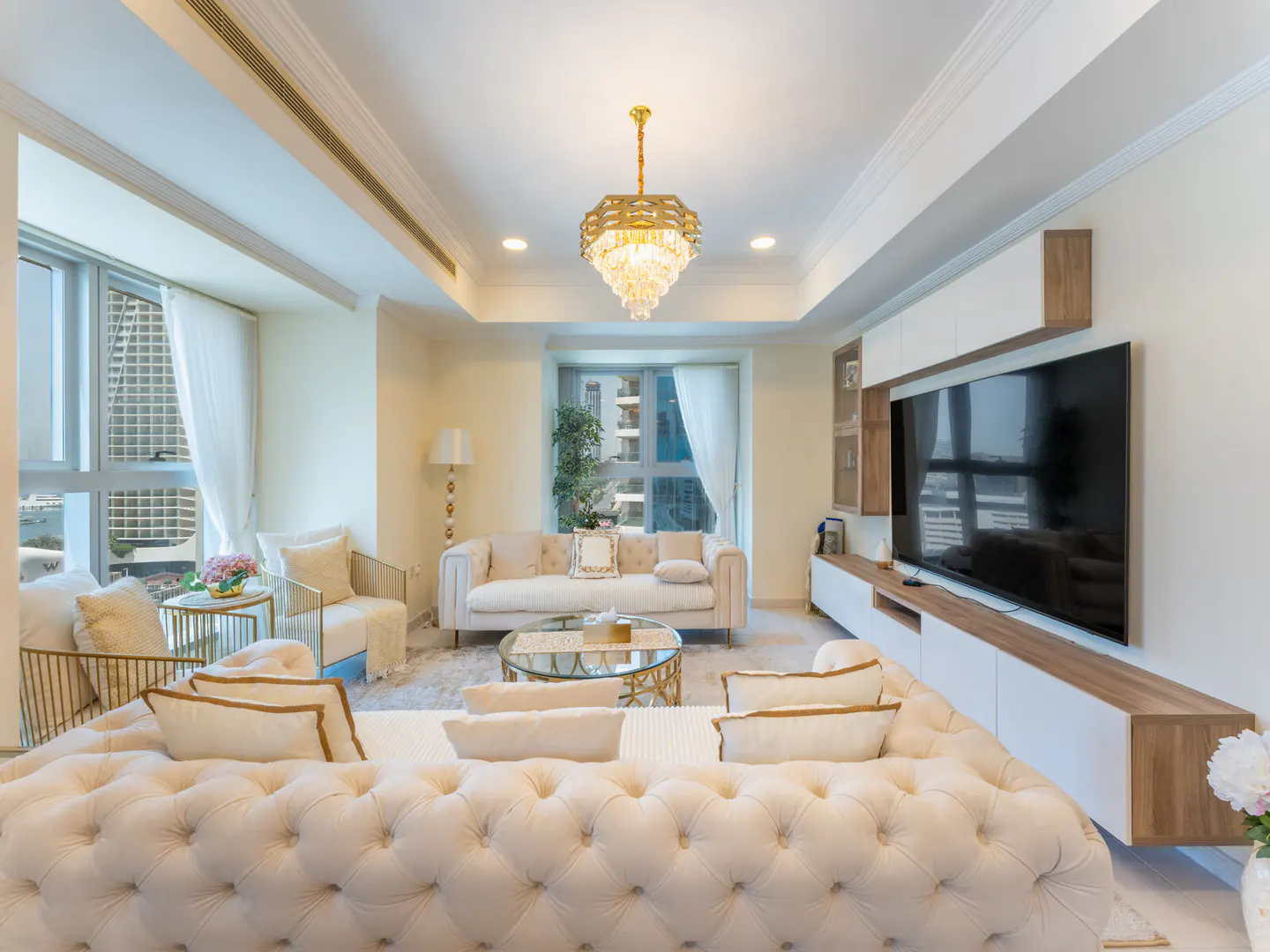 Bright living room with a tufted cream sofa, gold chandelier, and large windows with city views. A TV is mounted on a white and wood entertainment center.