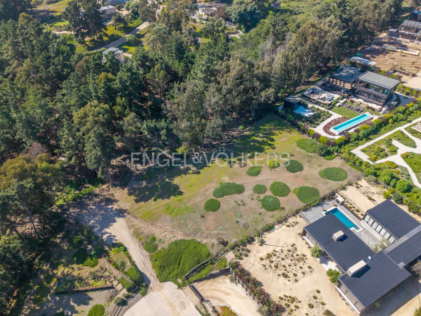 Aerial view of a grassy lot surrounded by trees and luxury homes with pools. Engel & Völkers logo is visible.