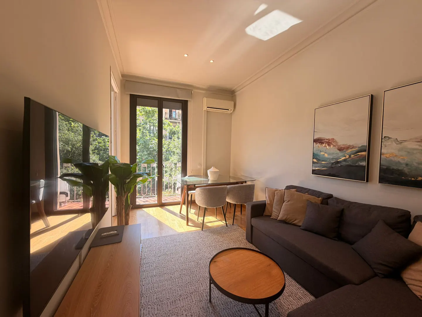Living room with gray sofa, wood coffee table, and dining table near balcony. Two landscape paintings hang on the wall.