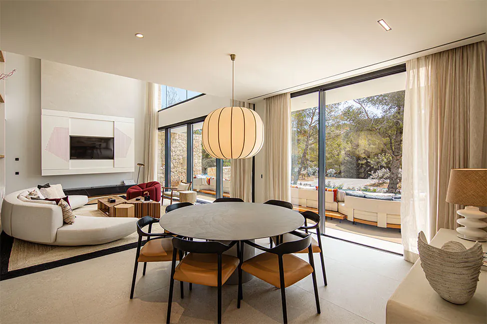 Bright, modern living room with a round dining table, curved sofa, and large windows overlooking a lush outdoor space.