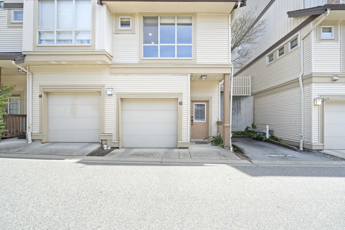 A two-story townhouse with beige siding, white garage doors, and a light brown front door.