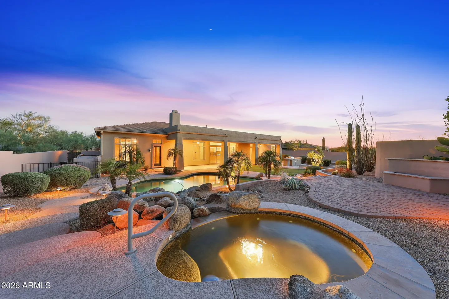 Backyard view of a tan house with a pool, spa, and desert landscaping at dusk.