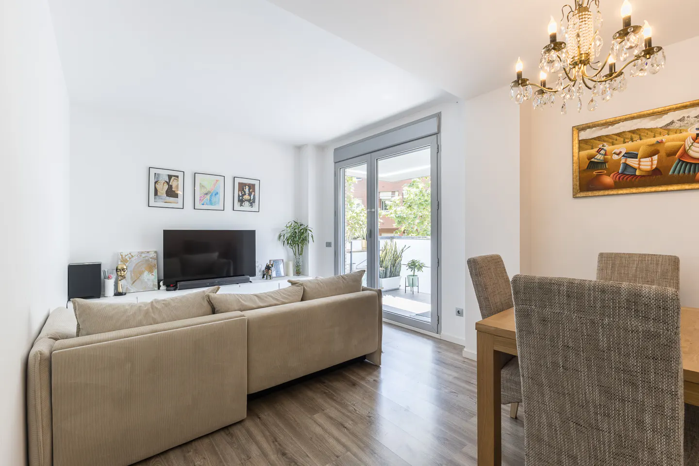 Bright living room with a beige sofa, TV, and dining table with a chandelier. Balcony doors let in natural light.