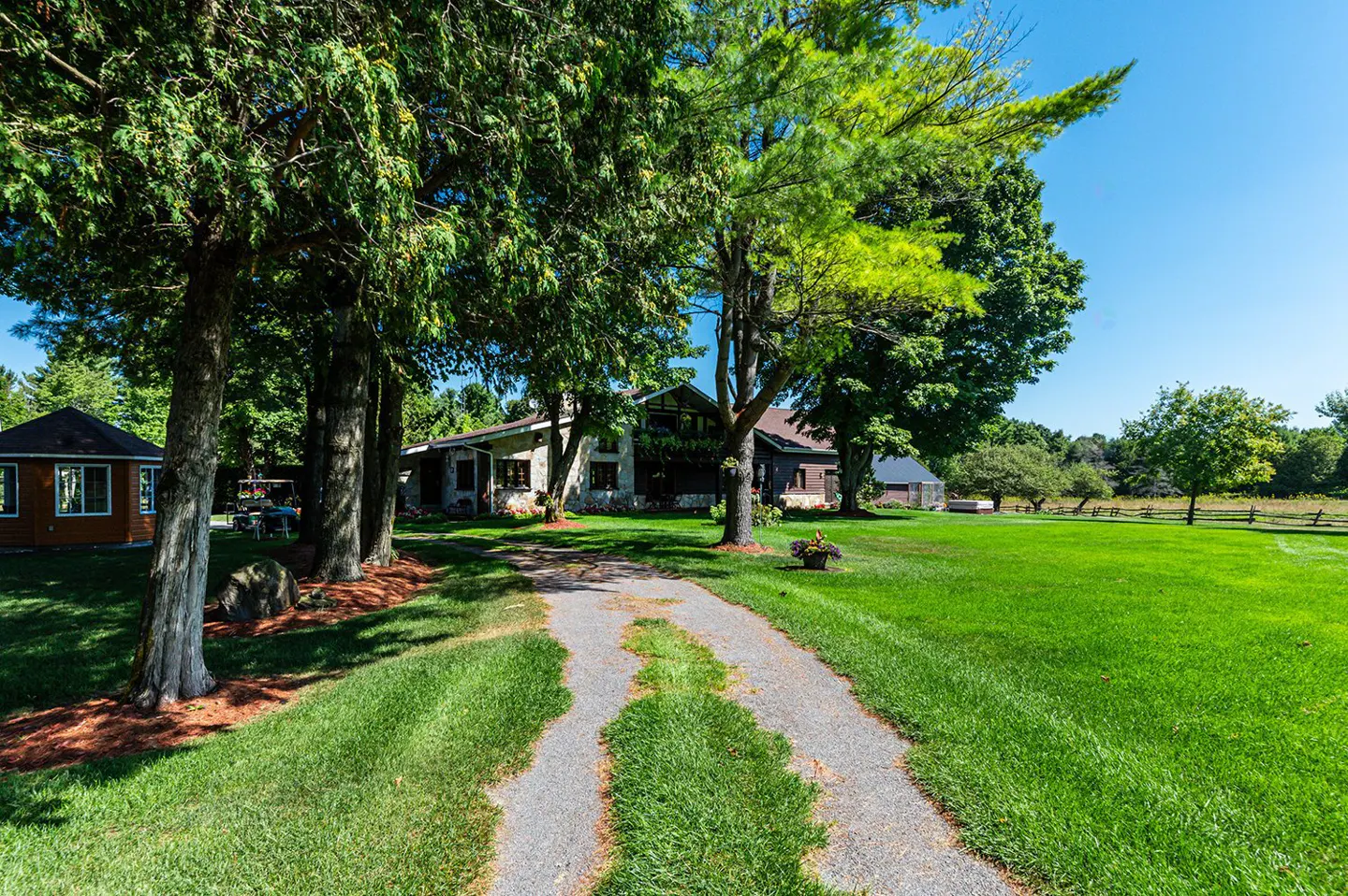Gravel driveway leads to a stone house surrounded by green lawn and trees under a blue sky. A small brown building is on the left.
