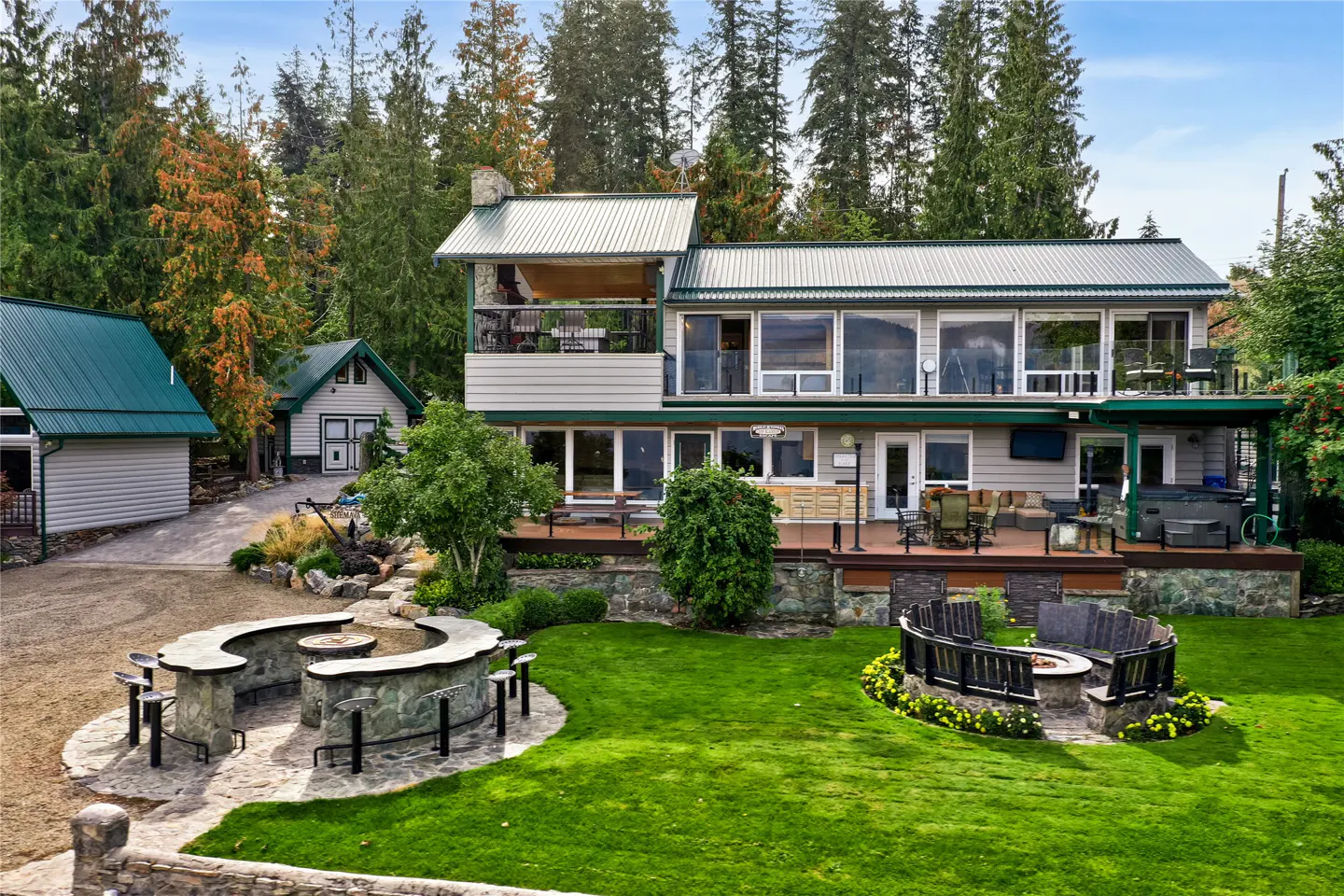 Two-story house with a green lawn, stone fire pits, and a deck. Tall trees surround the property.