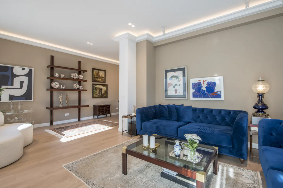 Living room with blue velvet sofa, glass coffee table, and wooden shelves displaying art and decorative objects.