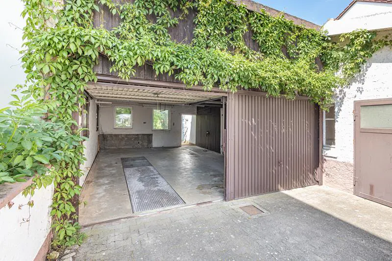 Open garage with a brown door, partially closed, and green vines growing on the roof and walls. The interior is visible with two windows.