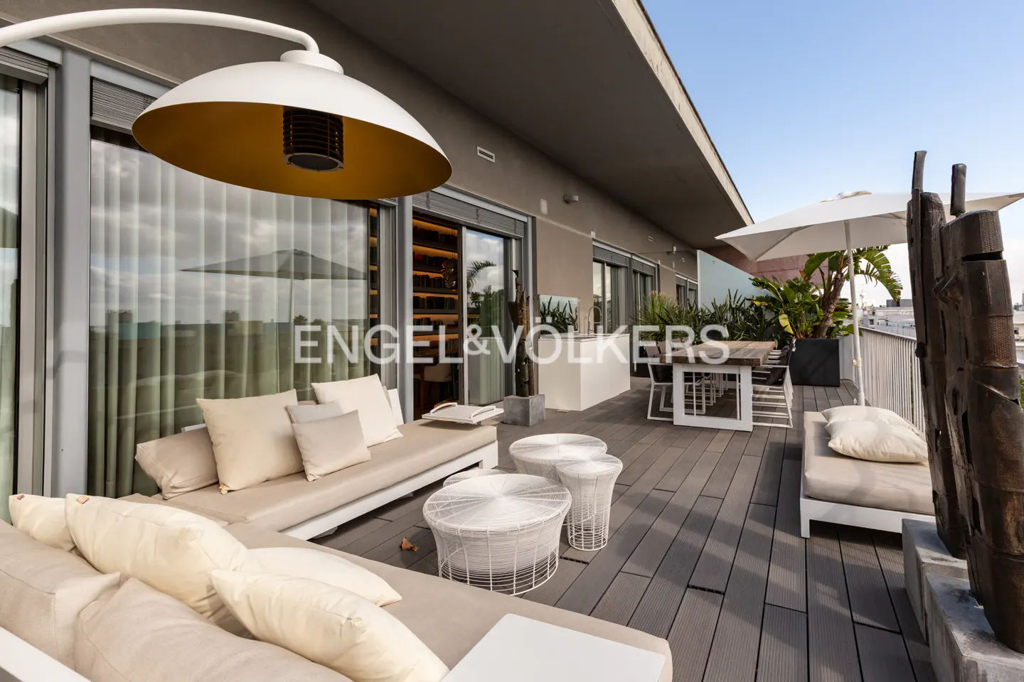 A modern rooftop terrace with a large white lamp, beige couches, white tables, and a dining area under a white umbrella.