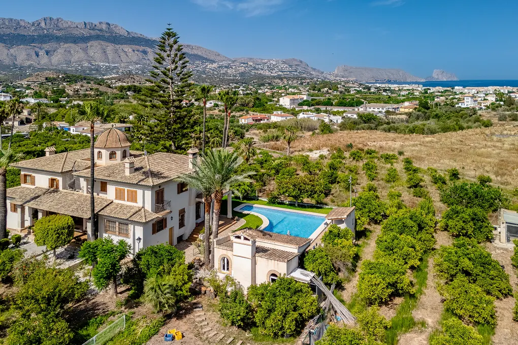 Aerial view of a large, beige villa with a pool, surrounded by green trees and mountains in the background under a blue sky.