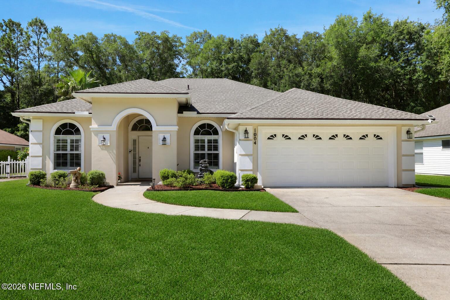 Single-story beige suburban home with a two-car garage, arched windows, and a manicured lawn against a backdrop of trees.