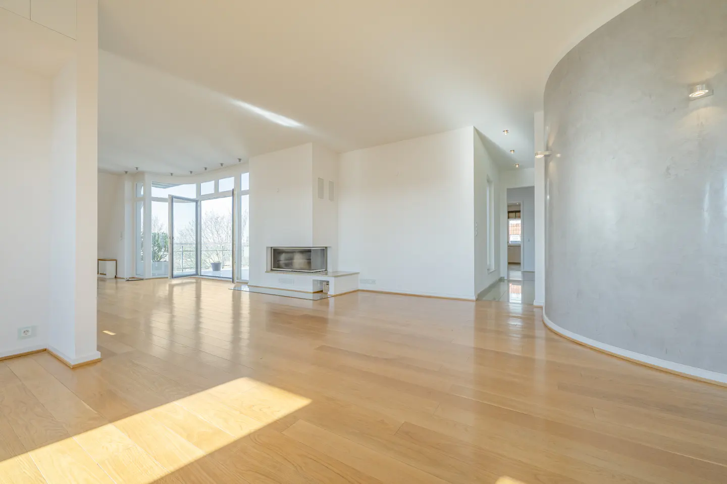 Bright, empty living room with light wood floors, white walls, fireplace, and curved wall. Balcony doors let in natural light.