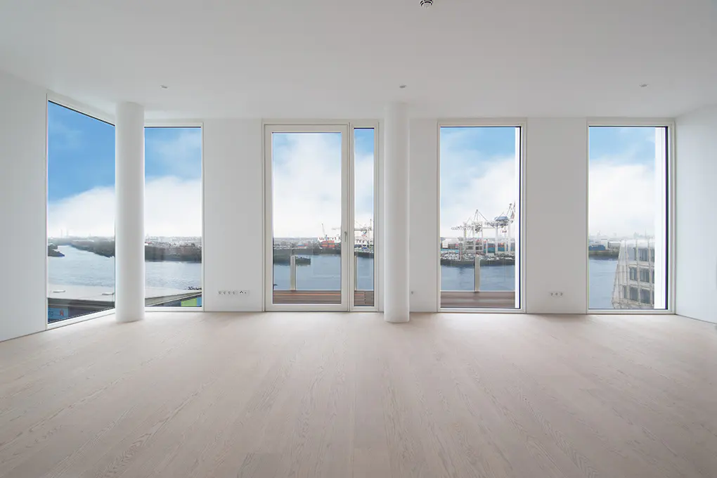 Bright, empty room with light wood floors and white walls. Large windows offer a harbor view with cranes and ships.