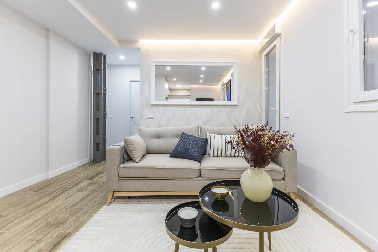 Bright living room with a beige sofa, decorative pillows, and a vase of dried flowers on black nesting tables. A mirror reflects the kitchen.