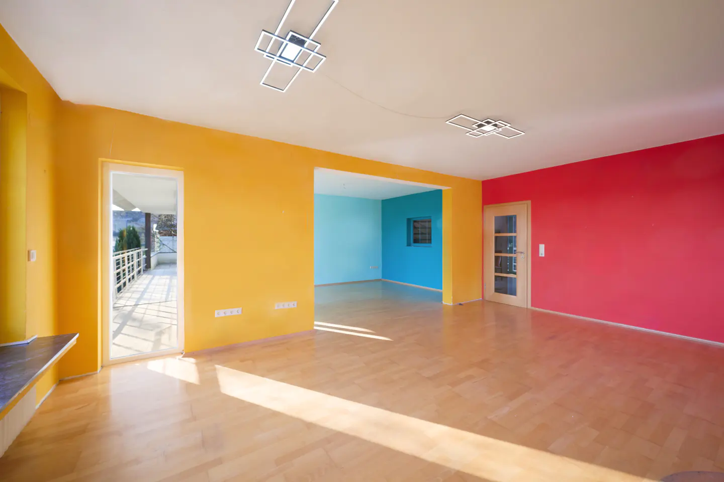 Bright, empty room with yellow, red, and blue walls, light wood floors, and modern light fixtures. A door leads to a balcony.
