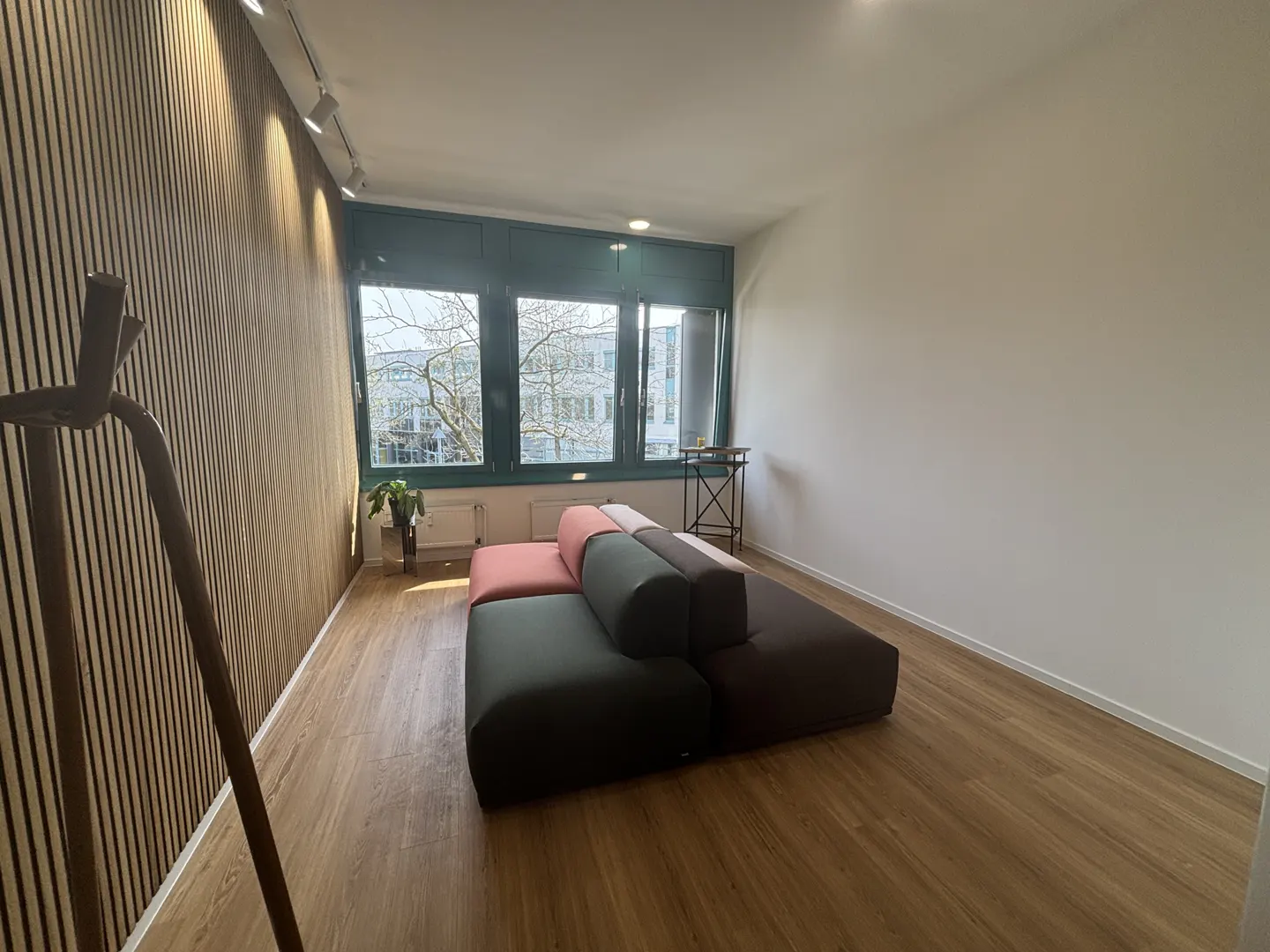 A modern living room with a modular sofa in shades of pink, green, and brown. Large windows offer natural light, and a wooden slat wall adds texture.