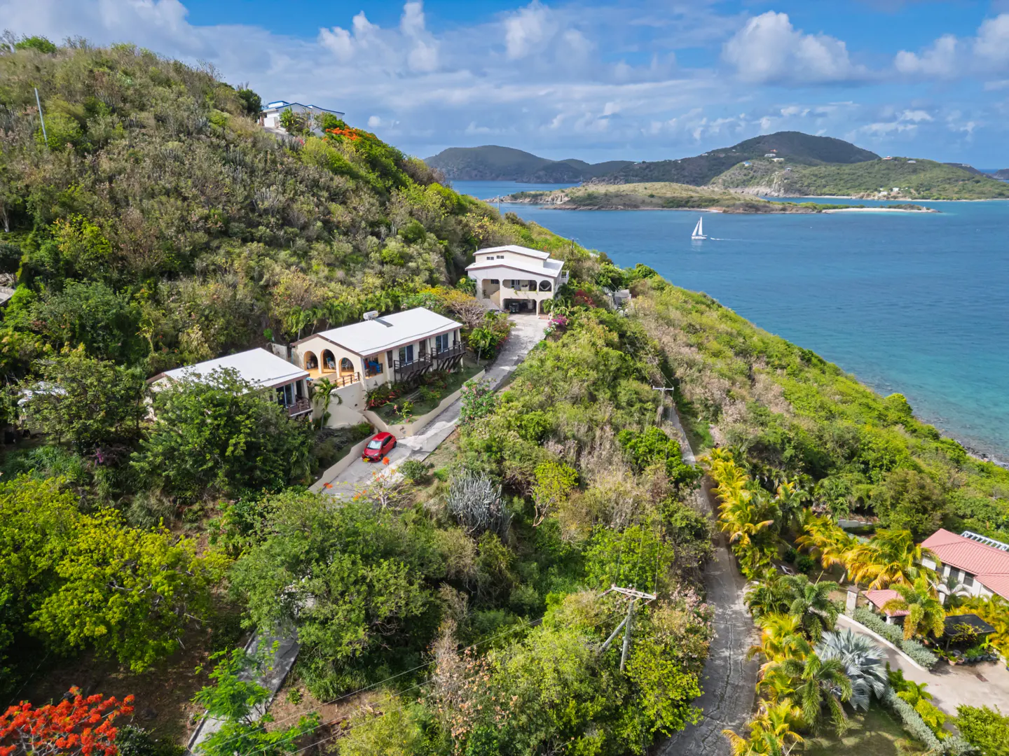 Aerial view of hillside homes with white roofs, a red car, and ocean views under a blue sky with scattered clouds.