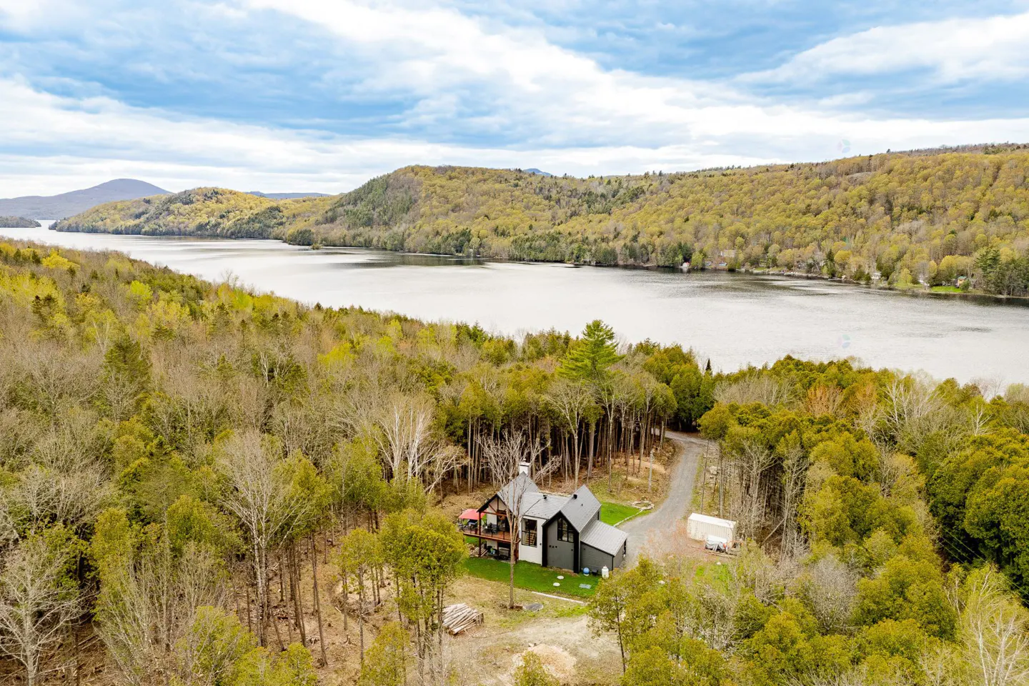 Aerial view of a modern dark gray house nestled in a forest near a lake, under a cloudy sky.